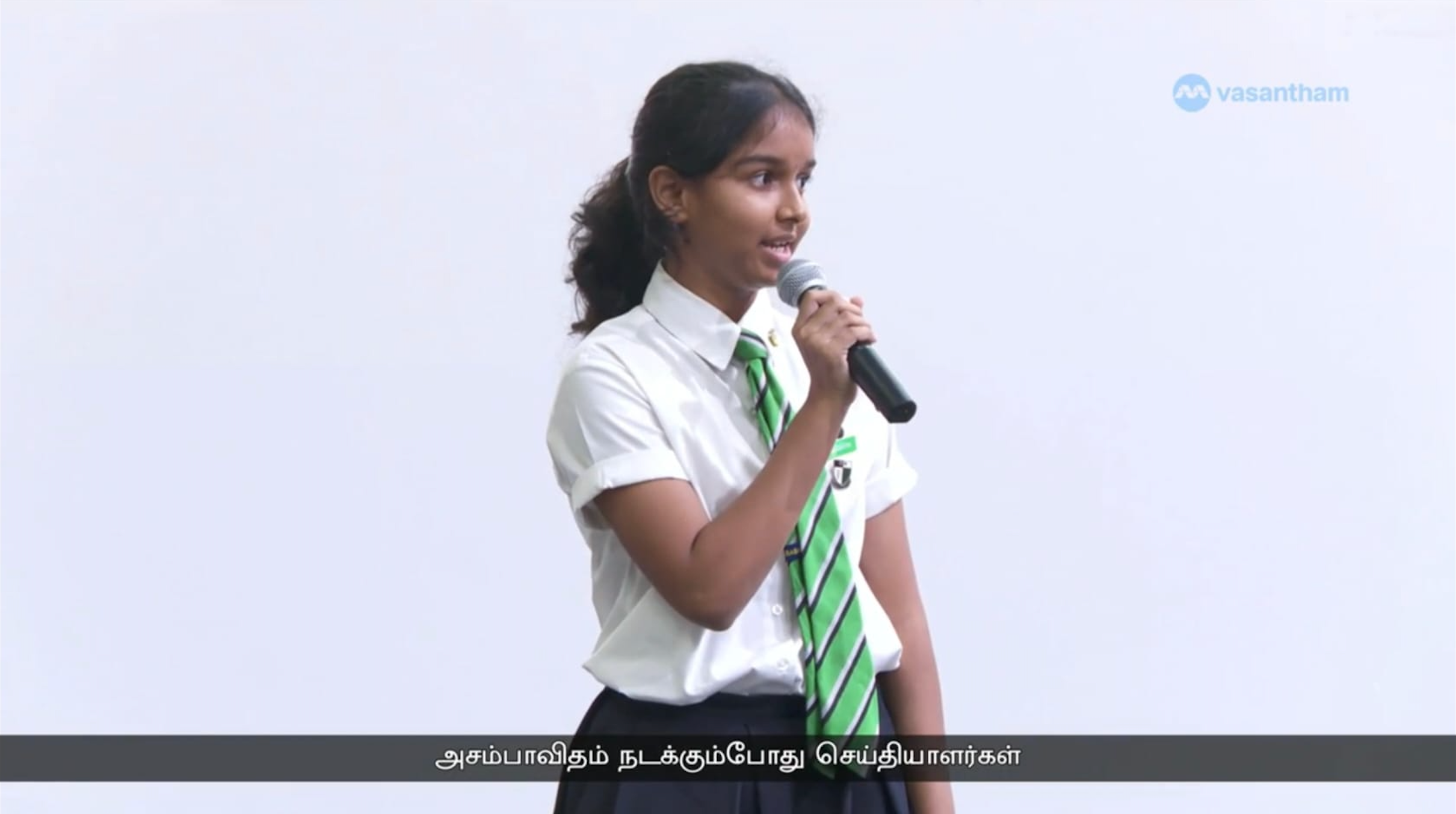 A screen capture of the Vasantham news telecast of a female student speaking with a microphone, looking into the audience.